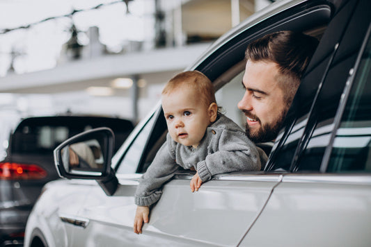 A smiling father looking at his baby, who is peeking out of an open car window