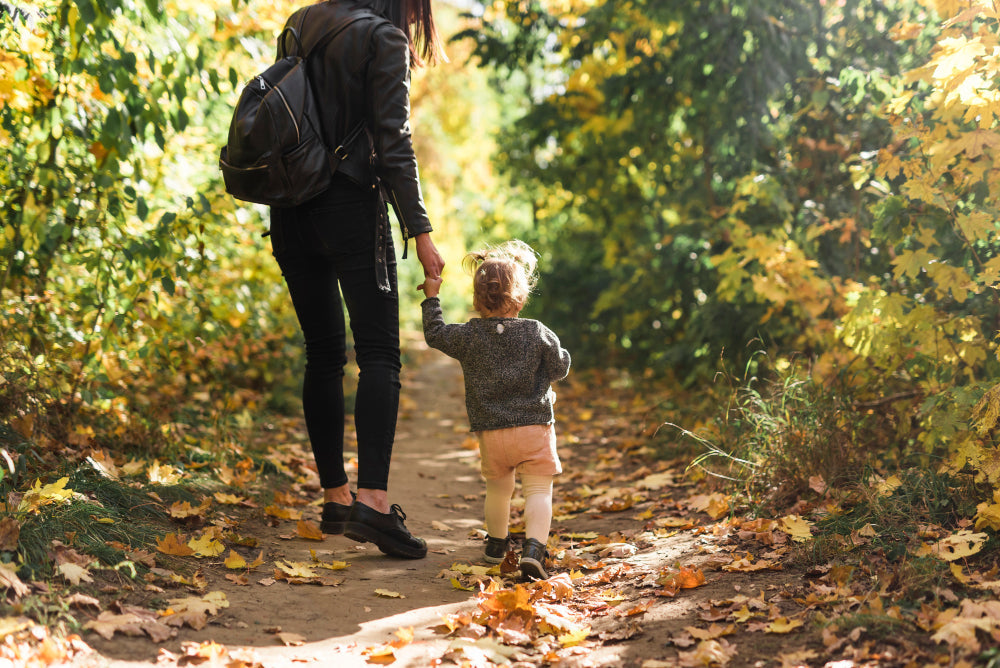 A mother and child walking hand-in-hand down a path covered with autumn leaves