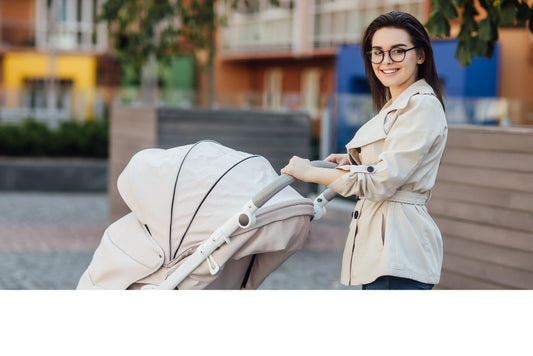 A woman wearing a beige trench coat pushing a baby stroller outdoors.