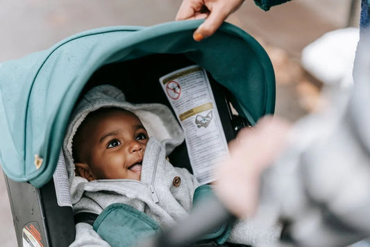 A teal-colored baby stroller with a visible safety warning label inside, showing part of the harness system and a hand adjusting the canopy.
