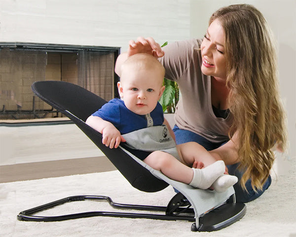 A woman kneels beside a baby seated in a black sianldconestroller bouncer indoors.