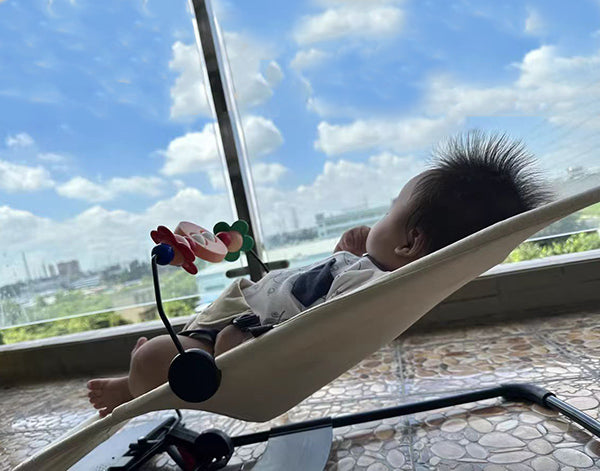 A baby is resting in a light-colored baby bouncer with a toy mobile, looking out at a cloudy blue sky and distant cityscape.