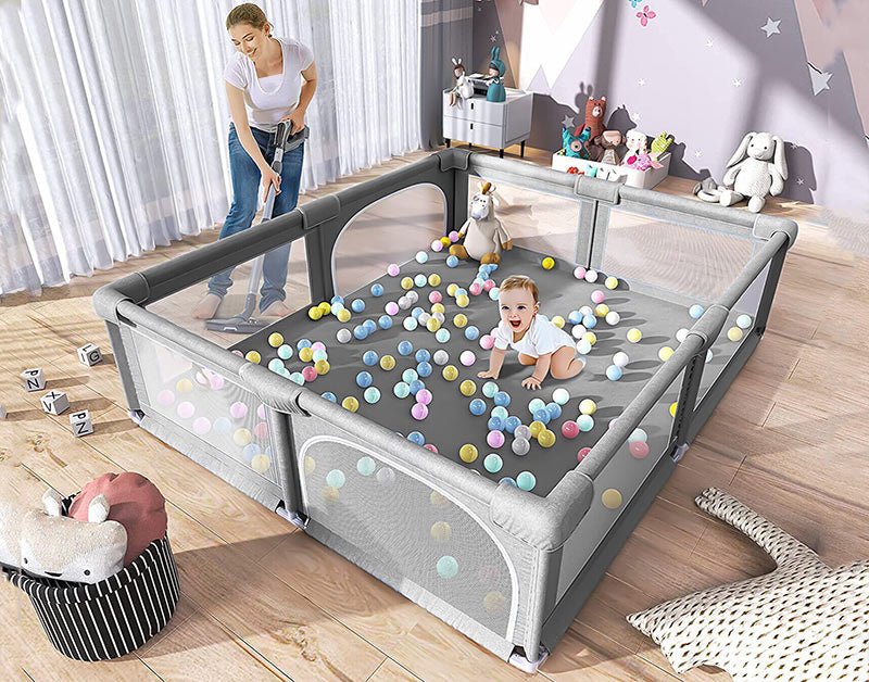  A woman vacuums around a smiling baby sitting in a large gray playpen filled with colorful ball pit balls.